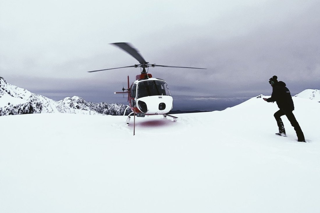 A helicopter on a remote snowy mountain summit with a person walking through the snow.